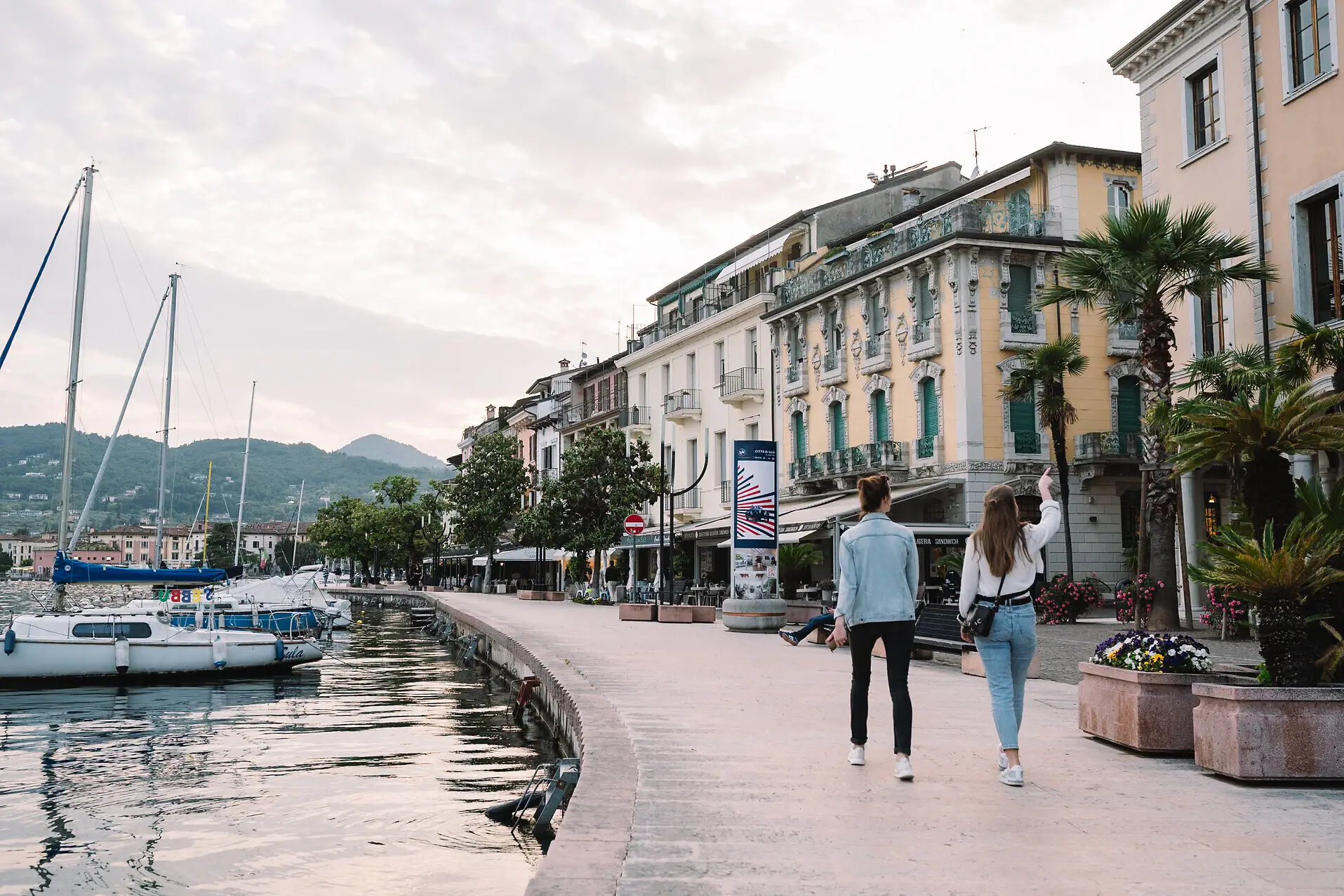 Two women walking on a sidewalk next to a body of water with boats and a ship in the background.