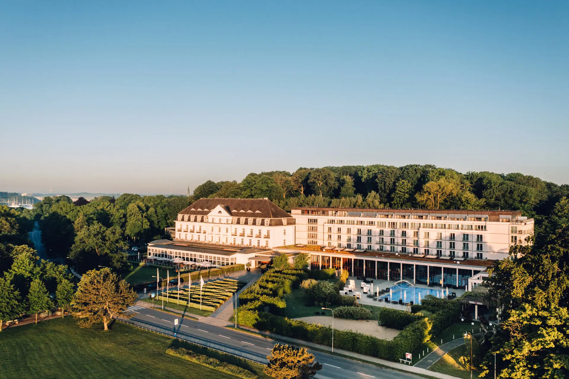 A-ROSA Travemünde Large white building with pool in the foreground and trees in the background.