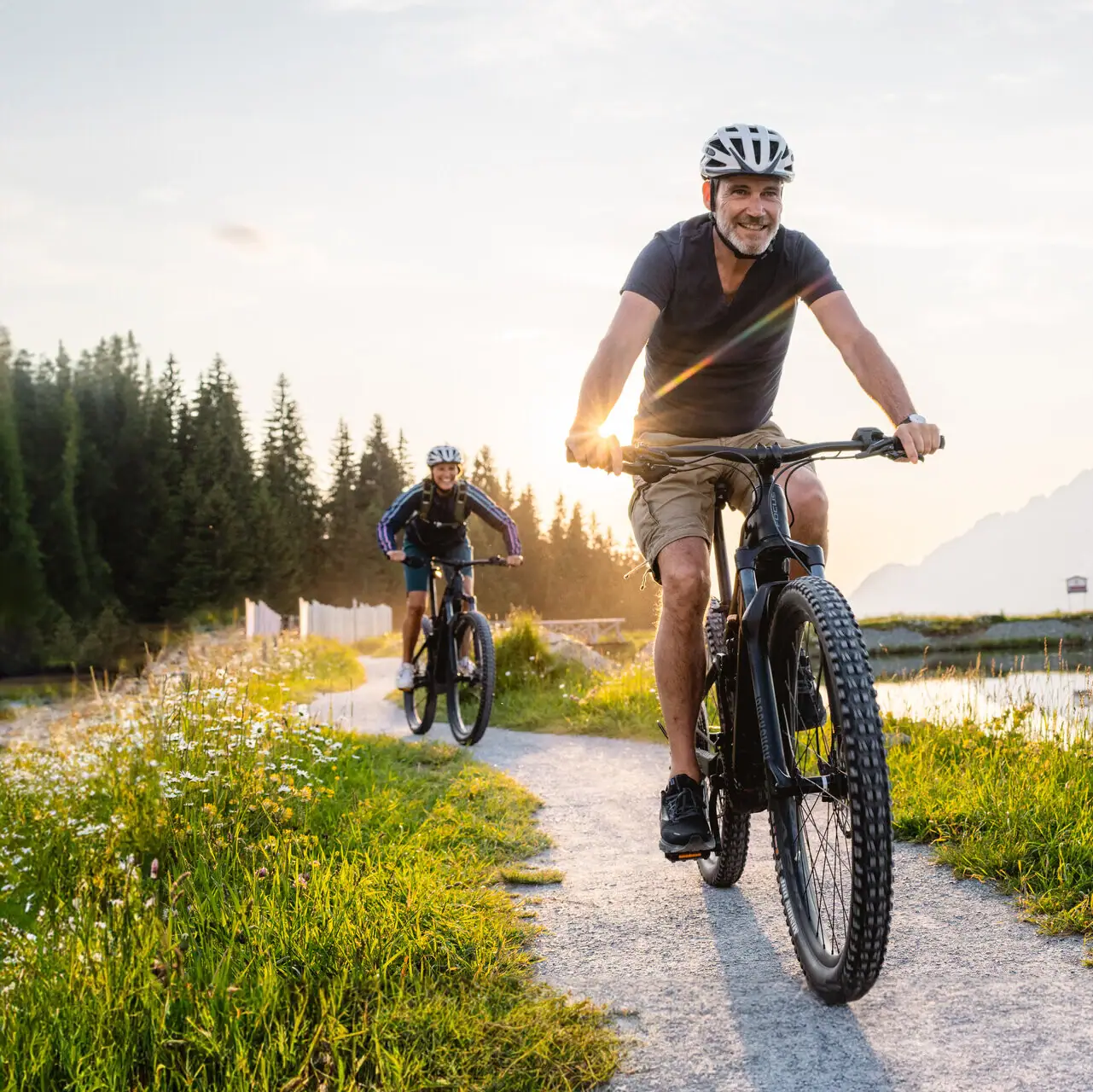 A man cycling on a path with a lake and mountains in the background.