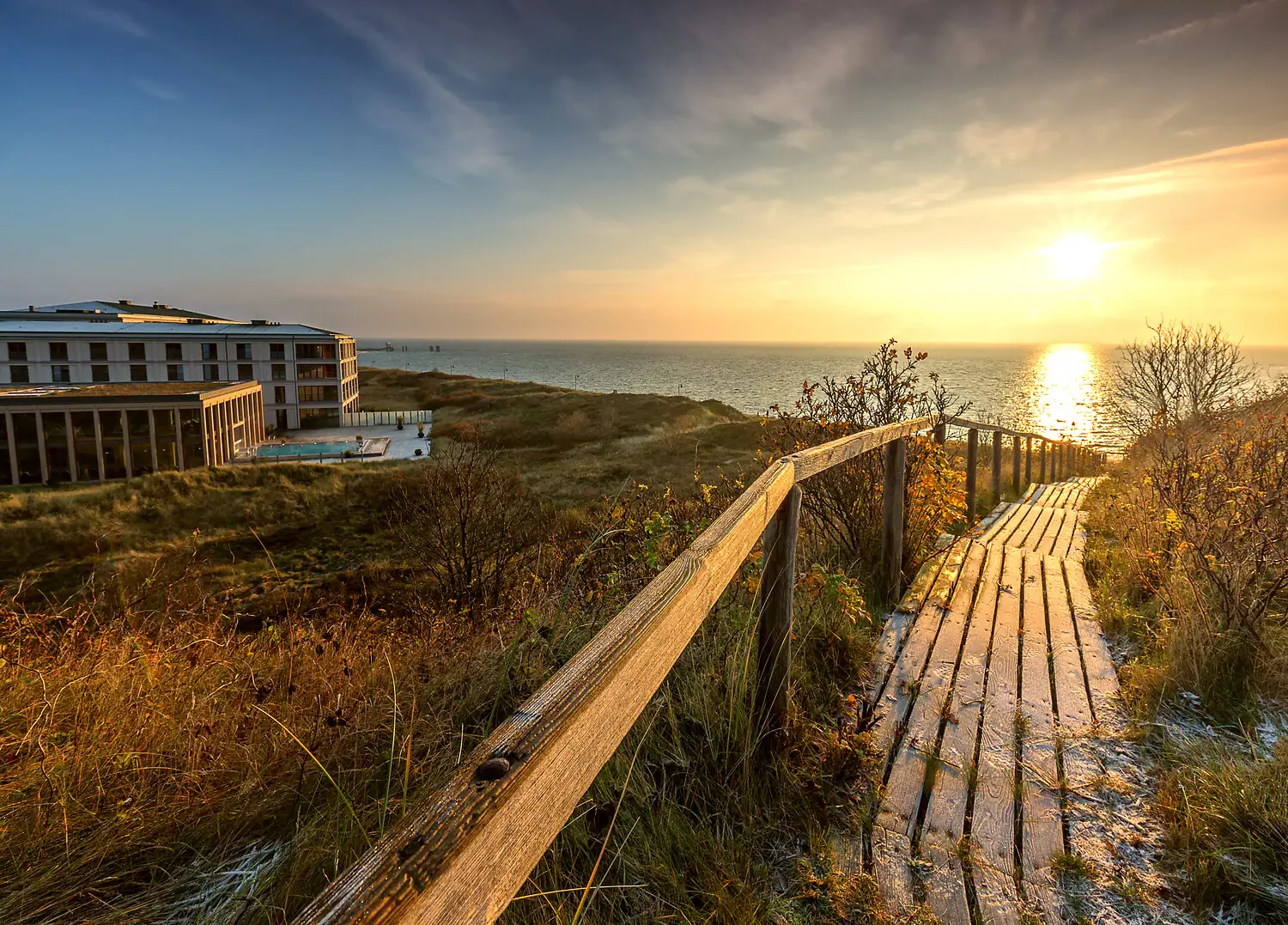 A-ROSA Sylt Wooden walkway leads to the beach with buildings and water in the background.
