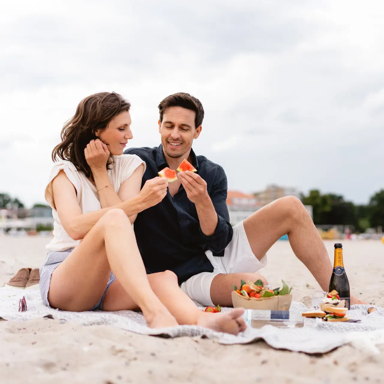 A man and woman sitting on a blanket on a beach enjoying a meal.
